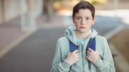 Ein Schüler trägt einen grauen Kapuzenpullover und hat einen Rucksack auf den Schultern. Er steht auf einem Gehweg im Freien.