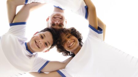 Drei Jungen in weißen Sporttrikots mit blauen Rändern stehen im Kreis und umarmen sich, lächeln in die Kamera