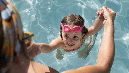 Kind mit Schwimmbrille hält Hände eines Erwachsenen im Schwimmbecken
