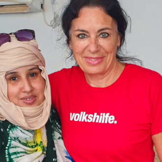 Zwei Frauen stehen nebeneinander und lächeln in die Kamera. Die Frau links trägt ein beiges Kopftuch, ein buntes Gewand in Grün, Weiß, Gelb und Blau. Die Frau rechts trägt ein rotes T-Shirt mit weißem Schriftzug „volkshilfe.“ und hat dunkle, zurückgebundene Haare. Sie legt den Arm um die andere Frau. Der Hintergrund ist neutral mit einer weißen Wand und einer Klimaanlage oben links im Bild.
