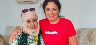 Zwei Frauen stehen nebeneinander und lächeln in die Kamera. Die Frau links trägt ein beiges Kopftuch, ein buntes Gewand in Grün, Weiß, Gelb und Blau. Die Frau rechts trägt ein rotes T-Shirt mit weißem Schriftzug „volkshilfe.“ und hat dunkle, zurückgebundene Haare. Sie legt den Arm um die andere Frau. Der Hintergrund ist neutral mit einer weißen Wand und einer Klimaanlage oben links im Bild.
