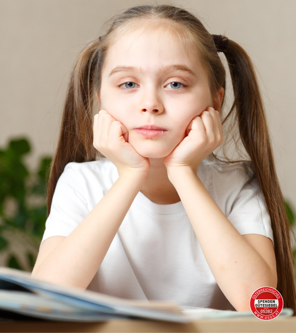 Ein Mädchen mit langen, geflochtenen Haaren sitzt mit den Händen im Gesicht auf einem Tisch. Im Vordergrund sind Bücher sichtbar. Im Bild: Logo Österreichisches Spendengütesiegel