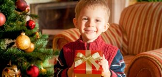 Kind in rotem Pullover hält Geschenk mit rotem Papier und goldener Schleife vor geschmücktem Weihnachtsbaum und gestreiftem Sofa