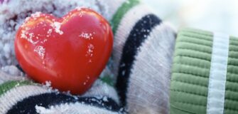 Zwei Hände mit grauen, grünen und weißen Handschuhen halten ein rotes Herz, das mit Schnee bedeckt ist