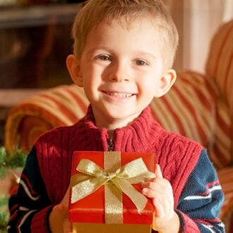 Bub trägt roten Pullover mit Zopfmuster und hält rotes Geschenk mit goldener Schleife vor sich
