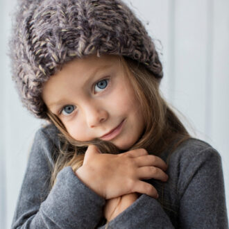 Kind mit grauer Strickmütze und grauem Pullover hält die Hände an die Brus
