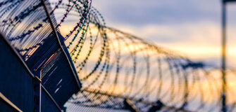 Stacheldraht auf einem hohen Metallzaun vor unscharfem Himmel bei Sonnenuntergang