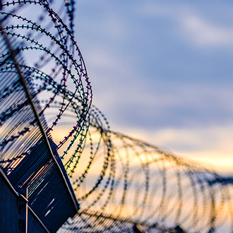 Stacheldraht auf einem hohen Metallzaun vor unscharfem Himmel bei Sonnenuntergang