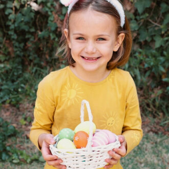 Herzensgeschenke Ostern - Mädchen steht im Garten und hält Körbchen gefüllt mir bunten Ostereiern