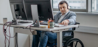 Ein Mann im Rollstuhl arbeitet in einem modernen Büro an einem höhenverstellbaren Schreibtisch mit zwei Monitoren und Headset.