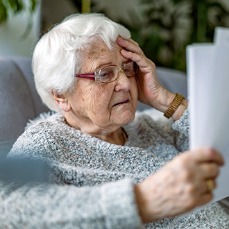 Ältere Frau sitzt auf einem Sofa und hält ein Dokument in der Hand, während sie sich nachdenklich an die Stirn fasst.