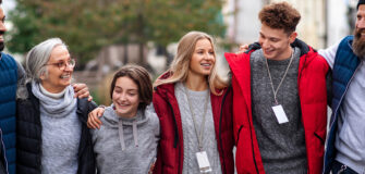 Fünf Menschen unterschiedlichen Alters gehen nebeneinander im Freien, lachen und haben die Arme umeinander gelegt.