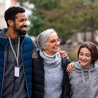 Drei Menschen unterschiedlichen Alters gehen nebeneinander im Freien, lachen und haben die Arme umeinander gelegt.