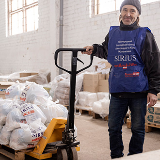 Ein Helfer in einer Lagerhalle in der Ukraine steht neben Paletten mit Lebensmitteln und Hilfsgütern im Rahmen der Ukraine-Hilfe und hält einen Hubwagen.