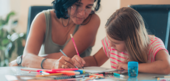 Frau sitzt mit einem Kind an einem Tisch und hilft beim Zeichnen oder Schreiben, auf dem Tisch liegen Stifte und Papier.