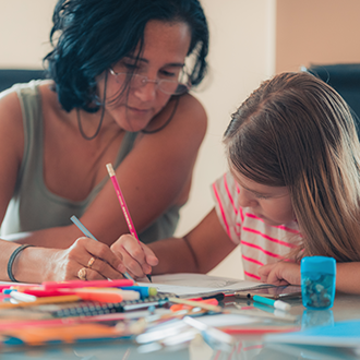 Frau sitzt mit einem Kind an einem Tisch und hilft beim Zeichnen oder Schreiben, auf dem Tisch liegen Stifte und Papier.