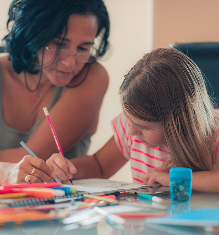Frau sitzt mit einem Kind an einem Tisch und hilft beim Zeichnen oder Schreiben, auf dem Tisch liegen Stifte und Papier.