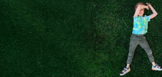 Ein Kind liegt in der Wiese, den linken Arm über dem Kopf, der rechte Arm liegt neben dem Kopf in der Wiese. Das Kind Blickt in die Kamera.