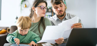 Eine Familie sitzt an einem Tisch und schaut gemeinsam auf Rechnungen, ein Kind malt daneben, ein Laptop steht auf dem Tisch.