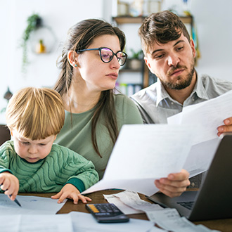Eine Familie sitzt an einem Tisch mit Unterlagen und Rechnungen, ein Kind zeichnet daneben.