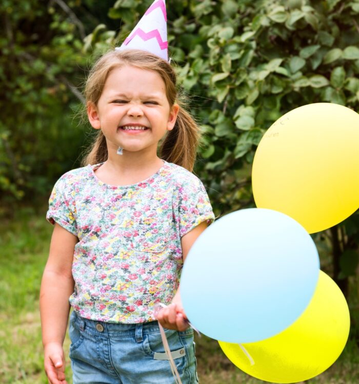 Kind mit geblümtem T-Shirt, Jeansshorts und Partyhut hält drei Luftballons in Gelb und Hellblau im Garten