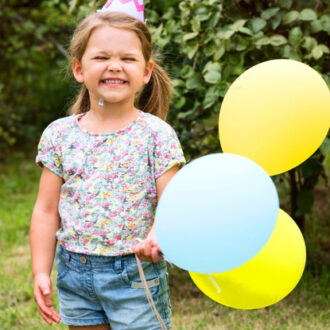 Kind mit geblümtem T-Shirt, Jeansshorts und Partyhut hält drei Luftballons in Gelb und Hellblau im Garten