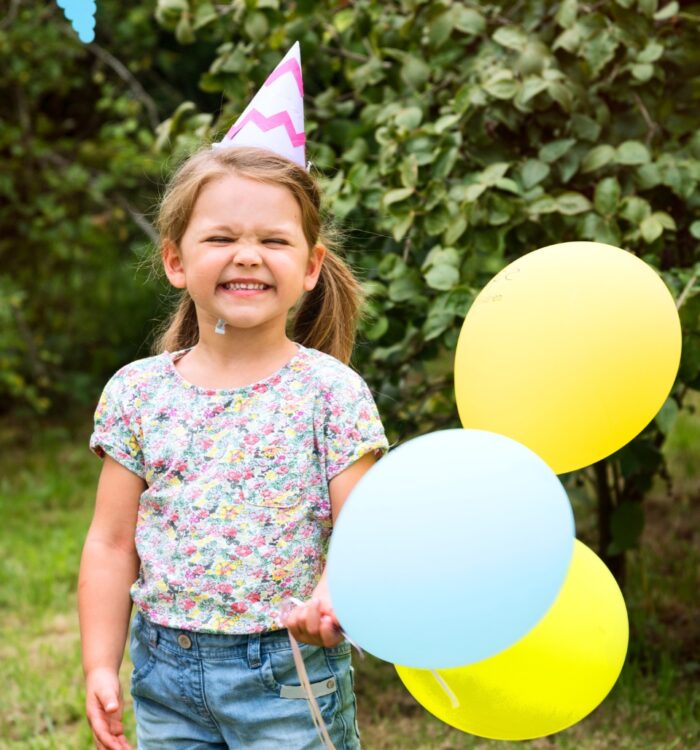 Kind mit geblümtem T-Shirt, Jeansshorts und Partyhut hält drei Luftballons in Gelb und Hellblau im Garten