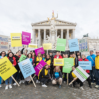 Mitarbeiter:innen von Caritas, Diakonie, Hilfswerk, Rotem Kreuz und Volkshilfe stehen mit Protestschildern vor dem österreichischen Parlament und fordern eine Pflegereform sowie bessere Rahmenbedingungen für Pflege und Langzeitpflege.