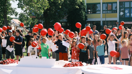 Schüler*innen der Volks- und Mittelschule St. Michael versammeln sich dem Schulgelände während einer Veranstaltung der Tour de Chance und winken mit roten Luftballons.