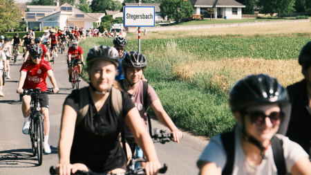 Ein langer Fahrradkonvoi fährt an einem Ortsschild von Stegersbach vorbei.