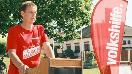 Hans Hager spricht beim Auftakt der Tour de Chance 2026 in Niederösterreich vor Teilnehmer*innen und Volkshilfe-Fahne.