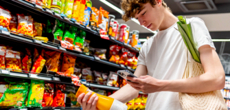 Ein junger Mann vergleicht in einem Supermarkt die Angaben auf einer Flasche mit seinem Smartphone. Er trägt einen Stoffbeutel mit Gemüse über der Schulter und steht vor einem Regal mit Lebensmitteln.