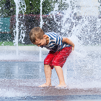 Ein Kind spielt an einem heißen Sommertag zwischen Wasserfontänen auf einem öffentlichen Platz.