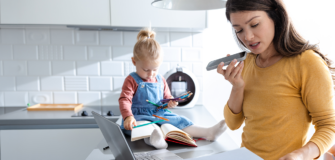 Eine Frau hält ein Smartphone in der Hand und blickt auf ein Blatt Papier. Auf der Kücheninsel steht ein Laptop. Im Hintergrund sitzt ein kleines Kind mit Stiften und einem offenen Buch auf der Arbeitsfläche.