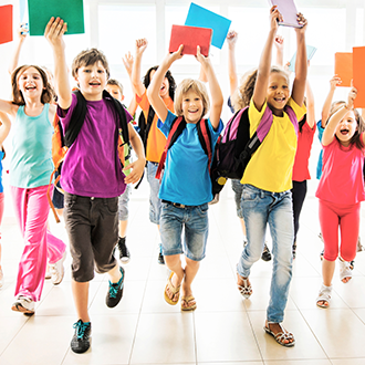 Eine Gruppe von Schulkindern läuft mit Schultaschen und bunten Heften durch einen hellen Raum auf die Kamera zu.