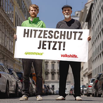 Marc Dengler von Greenpeace Österreich und Volkshilfe-Direktor Erich Fenninger halten auf einer Straße ein Transparent mit der Aufschrift „HITZESCHUTZ JETZT!“.
