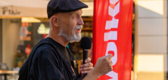 Erich Fenninger spricht bei der Sozialhilfe-Kundgebung in Klagenfurt mit einem Mikrofon vor einer roten Volkshilfe-Fahne.