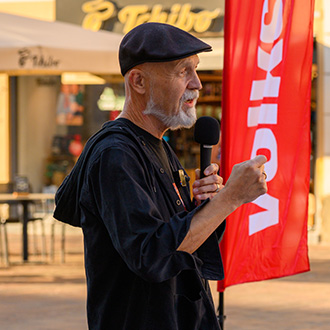 Erich Fenninger spricht bei der Sozialhilfe-Kundgebung in Klagenfurt mit einem Mikrofon vor einer roten Volkshilfe-Fahne.