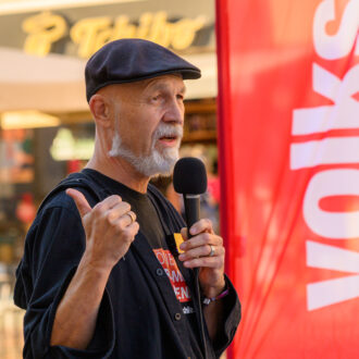 Erich Fenninger spricht bei der Kundgebung in Klagenfurt ins Mikrofon und gestikuliert neben einer roten Volkshilfe-Fahne.
