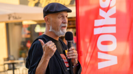 Erich Fenninger spricht bei der Kundgebung in Klagenfurt ins Mikrofon und gestikuliert neben einer roten Volkshilfe-Fahne.