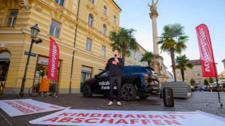 Erich Fenninger spricht zwischen Volkshilfe-Fahnen am Alten Platz in Klagenfurt. Vor ihm liegt ein Transparent mit der Aufschrift „Kinderarmut abschaffen“.