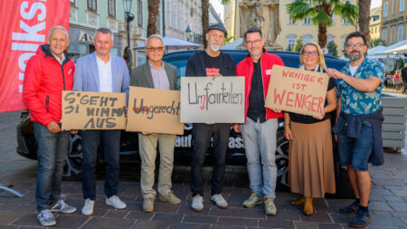 Erich Fenninger, Ernst Sandriesser und weitere Teilnehmer*innen halten in Klagenfurt Protestschilder, darunter „Weniger ist weniger“.