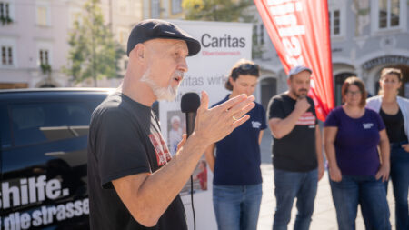 Erich Fenninger spricht in Linz ins Mikrofon und gestikuliert. Im Hintergrund stehen Teilnehmer*innen, ein Caritas-Aufsteller und eine Volkshilfe-Fahne.