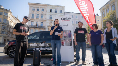Marion Huber spricht in Linz mit Mikrofon und Notizen vor einem Caritas-Aufsteller. Neben ihr stehen Erich Fenninger und weitere Teilnehmer*innen.
