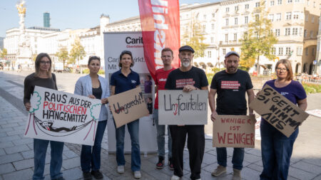 Marion Huber, Erich Fenninger und weitere Teilnehmer*innen halten am Linzer Hauptplatz Protestschilder, darunter „Weniger ist weniger“.