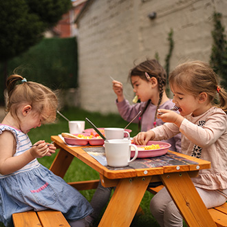 Drei Kinder sitzen an einem Holztisch im Garten und essen gemeinsam.